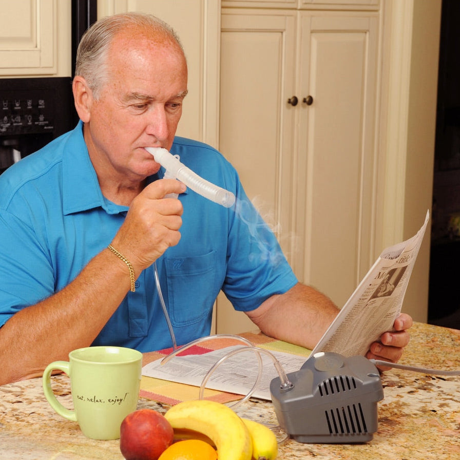 An older man in a blue shirt sits at a kitchen counter using a nebulizer with a tube connected to a gray machine, while reading a newspaper. 