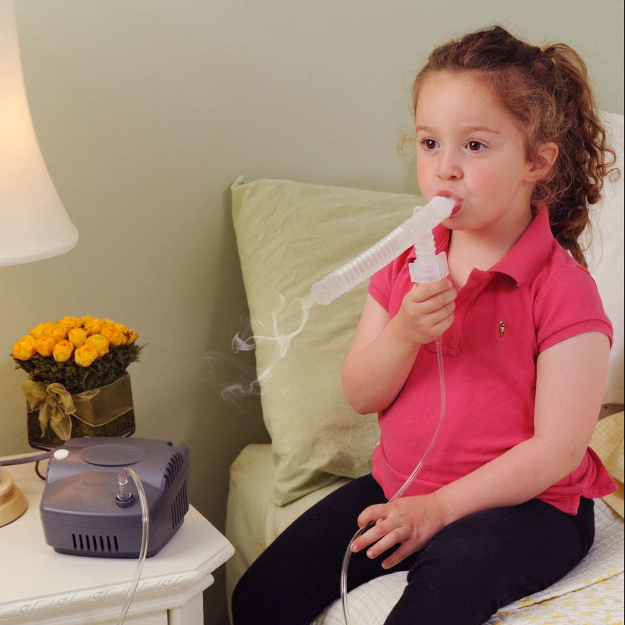 A young girl wearing a pink polo shirt sits on a bed using a nebulizer with a tube connected to a gray machine on a bedside table. A lamp, yellow flowers, and pillows are visible in the bedroom setting.