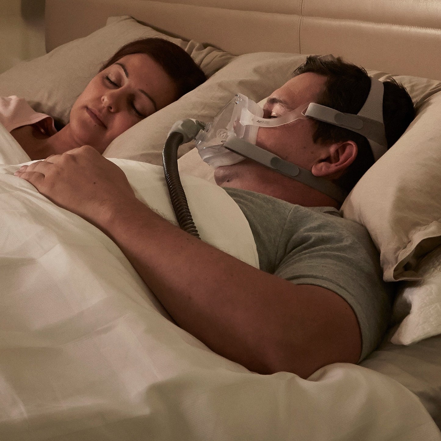 Man using a CPAP machine while sleeping next to a woman in bed.