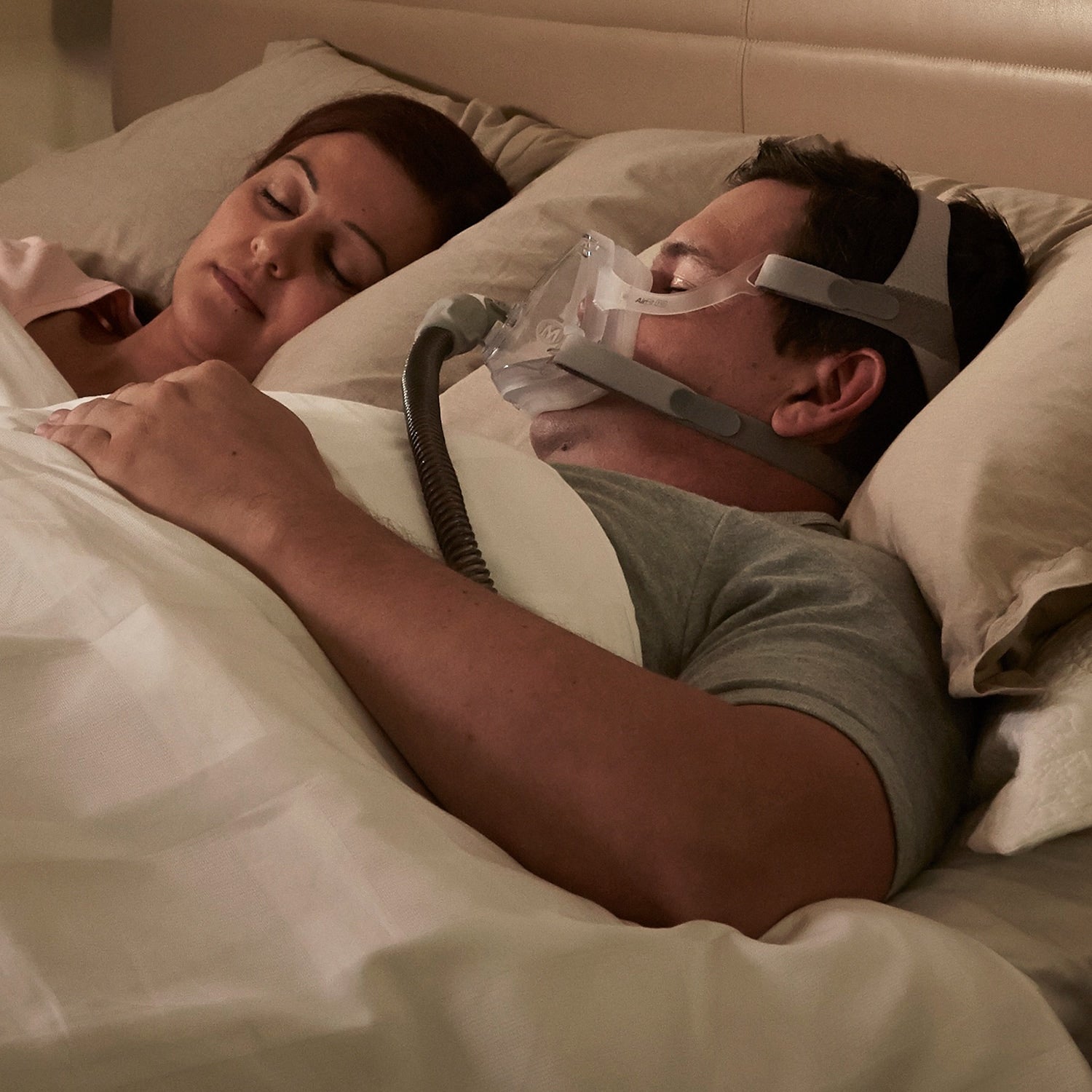 Man using a CPAP machine while sleeping next to a woman in bed.