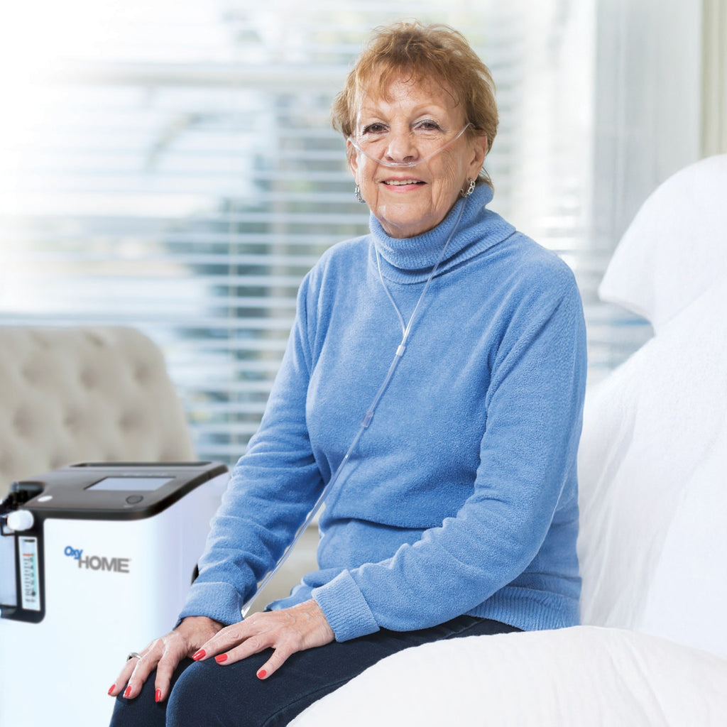 Woman in a blue sweater sitting on a couch, and wearing an oxygen nasal cannula, with an OxyHome Oxygen Concentrator in the background.