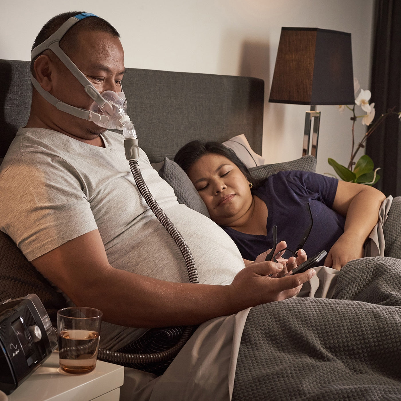 Man using a CPAP machine with a woman lying beside him in a bedroom setting.
