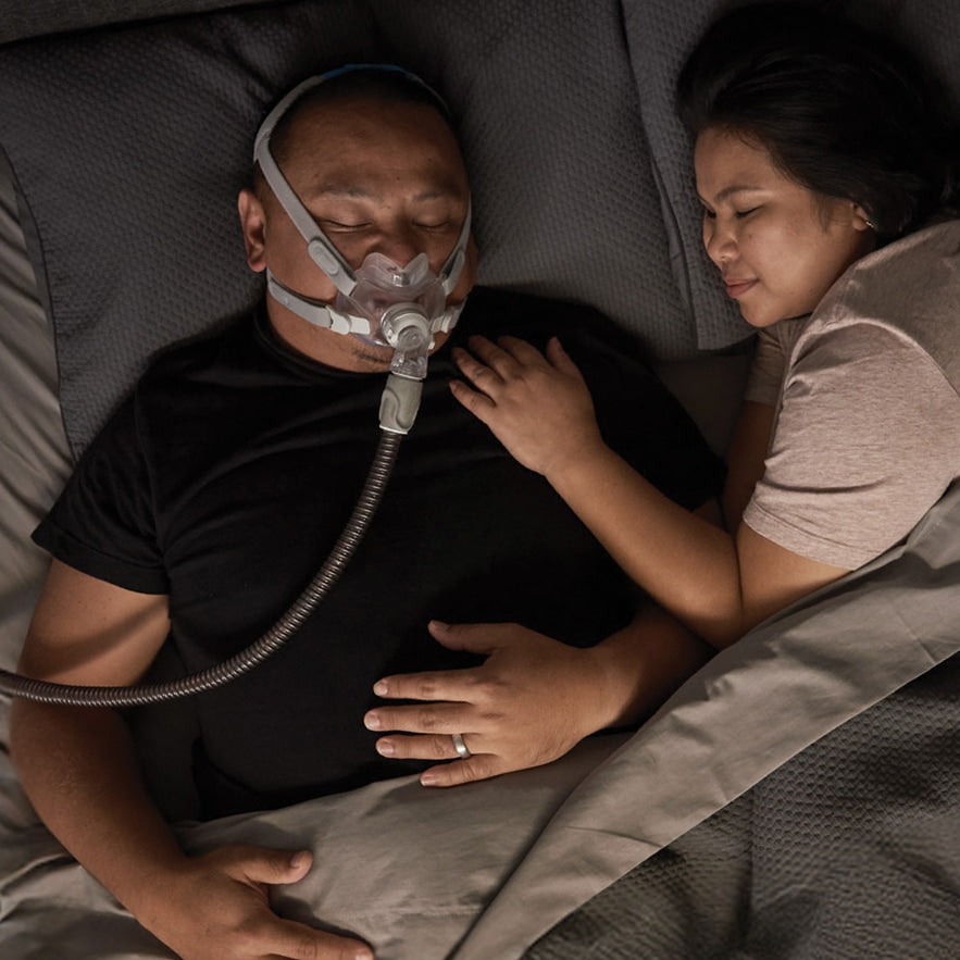 Man using a CPAP machine in bed with a woman beside him